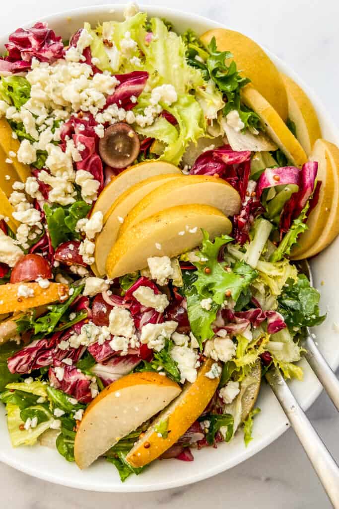 A closeup shot of an pear and blue cheese salad in a white serving bowl with silver serving spoons.