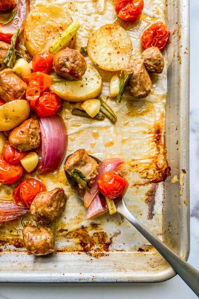 A closeup shot of the corner of a sheet pan with sausage, potatoes, and veggies with a silver spoon.