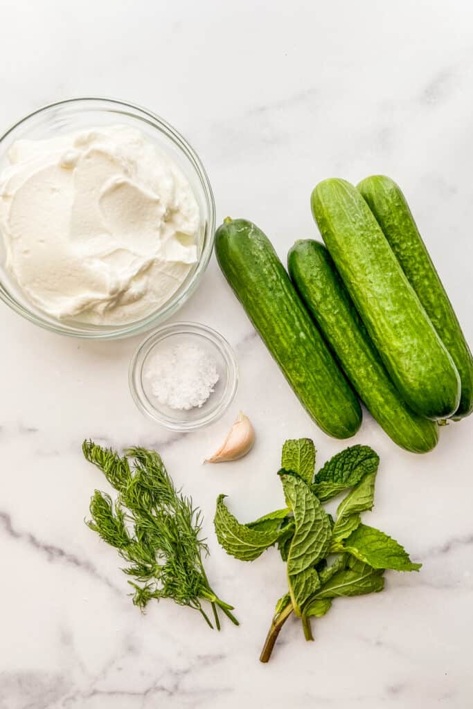 A bowl of Greek yogurt, four Persian cucumbers, mint, dill, sea salt, and a clove of garlic on a marble background.