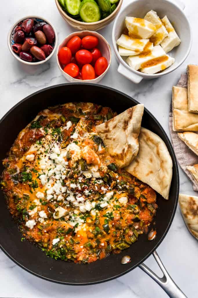 A skillet with menemen, next to some sliced pita, cheese, olives, cherry tomatoes, and sliced cucumber.