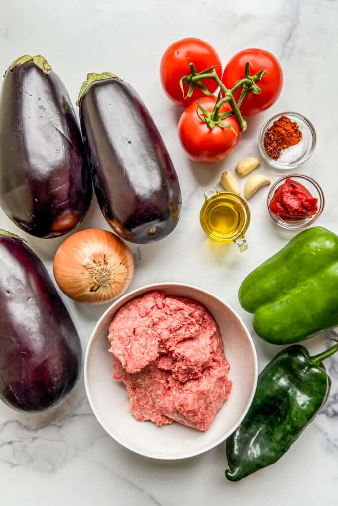 Eggplants, ground beef, a yellow onion, tomatoes, green peppers, tomato paste, garlic cloves, spices, and olive oil on a marble background.