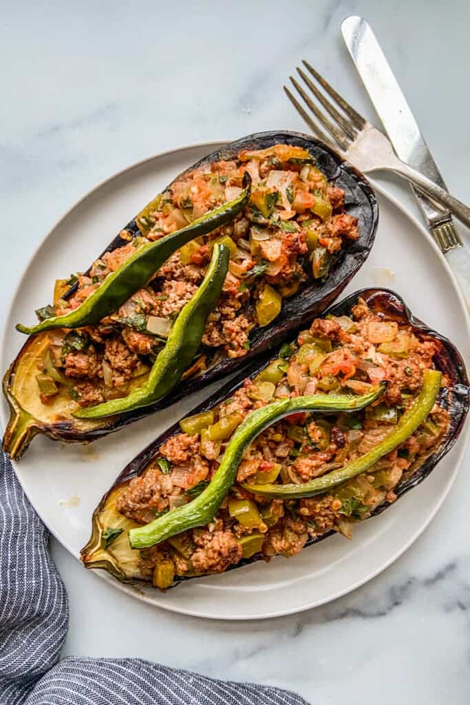 Two stuffed eggplants on a white plate, next to a fork and knife.