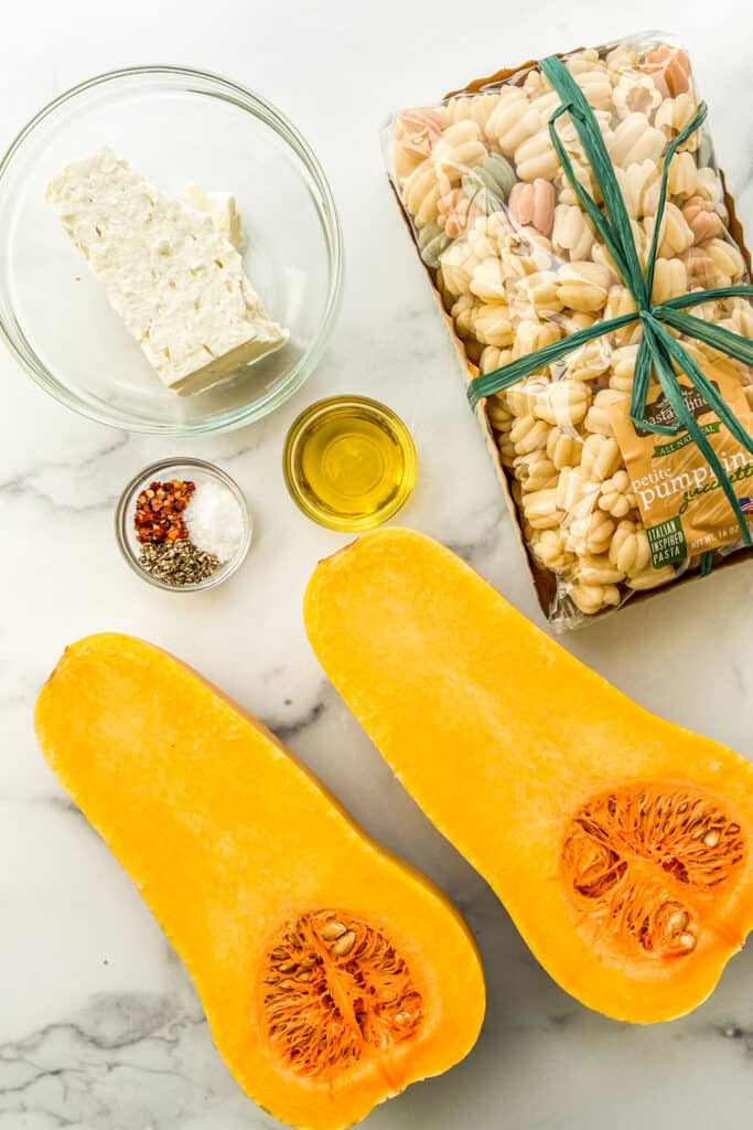 A halved butternut squash, a package of pasta, feta, olive oil, and spices.