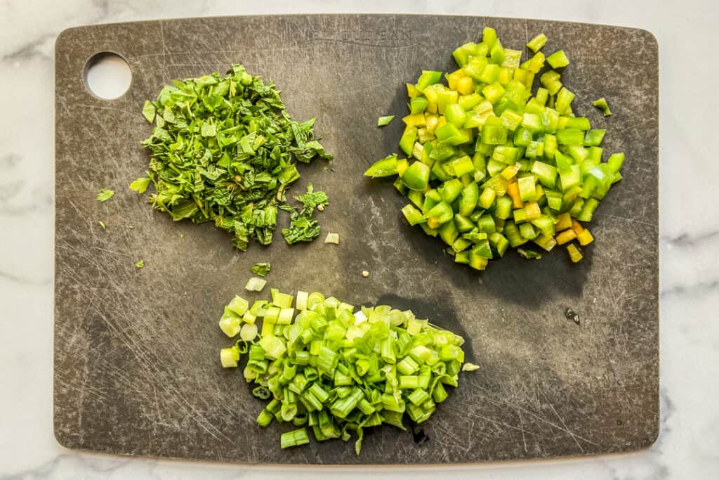 Diced bell pepper, herbs, and green onions on a black cutting board.