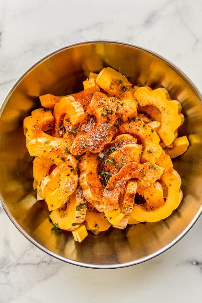 Chopped delicata squash tossed with spices and oil in a large metal mixing bowl.