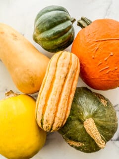 Types of winter squash stacked on each other - including delicata, kabocha, butternut, acorn, pumpkin, and spaghetti squash.