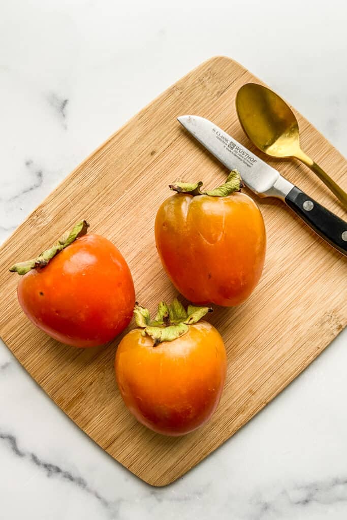 Three persimmons on a small, wooden cutting board with a gold spoon and small paring knife.