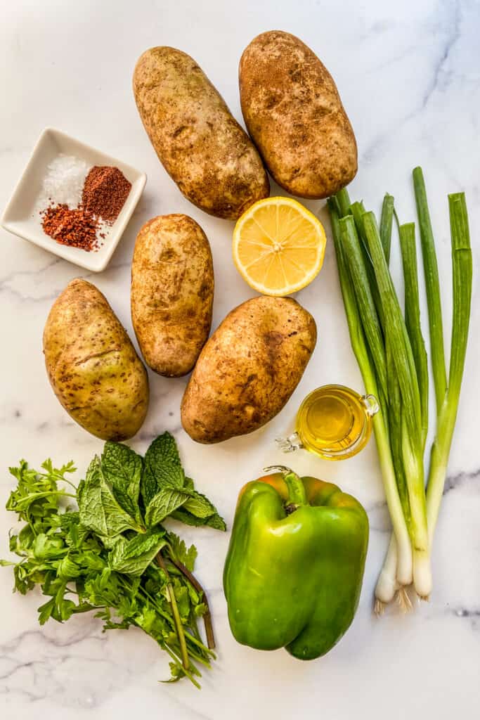 Potatoes, green onions, green pepper, herbs, half a lemon, olive oil, and spices on a marble background.