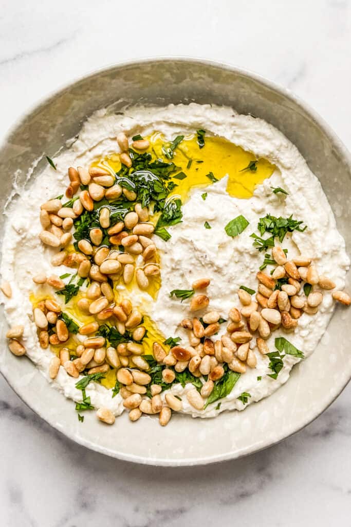 An overhead shot of a bowl of whipped feta dip topped with toasted pine nuts, olive oil, and chopped parsley.