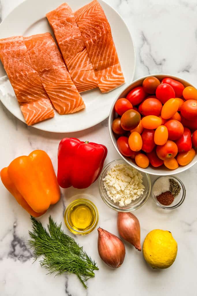 Salmon fillets, cherry tomatoes, bell peppers, shallots, feta, spices, oil, dill, and a lemon on a marble background.