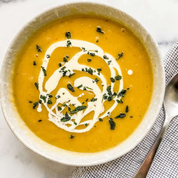 An overhead shot of a bowl of acorn squash soup with a cream drizzle, crumbled sage leaves, and a silver spoon.