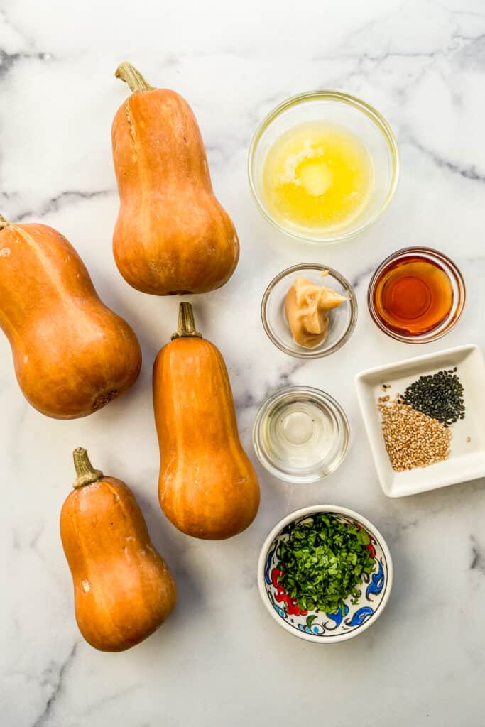 Four honeynut squash, melted butter, miso, maple syrup, rice vinegar, sesame seeds, and chopped cilantro on a marble background.