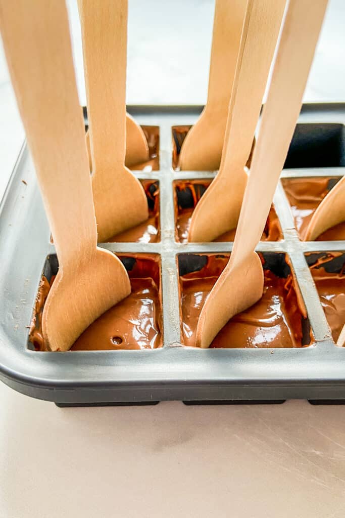 Melted chocolate in an ice cube tray with wooden spoons sticking out.