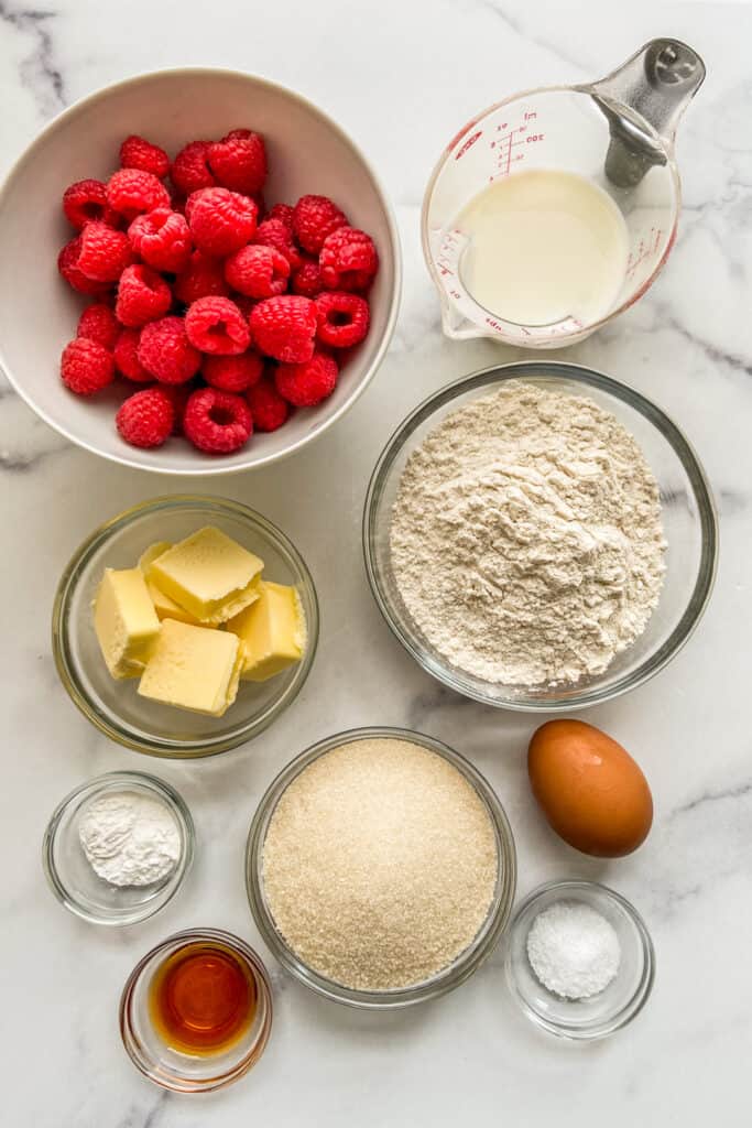Raspberries, milk, flour, sugar, butter, egg, baking powder, vanilla extract, and salt on a marble background.