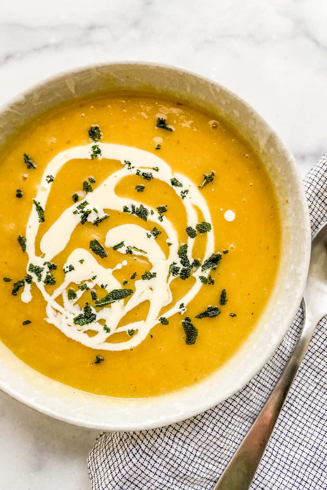 An overhead shot of a bowl of acorn squash soup with a cream drizzle, crumbled sage leaves, and a silver spoon.