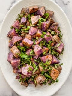 Roasted purple potatoes on a white platter topped with fresh herbs.
