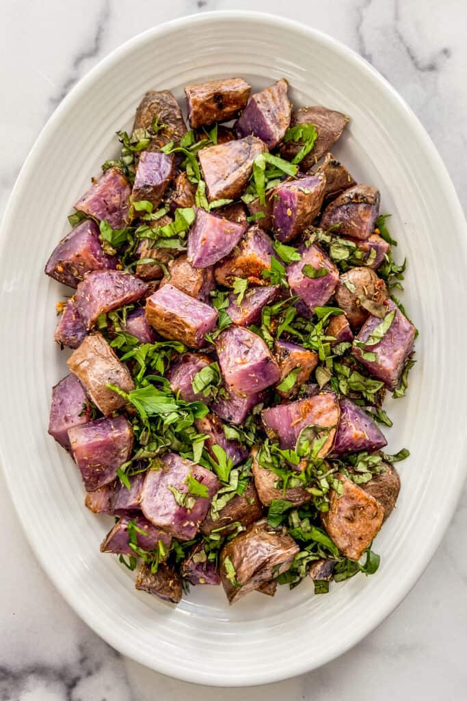 Roasted purple potatoes on a white platter topped with fresh herbs.
