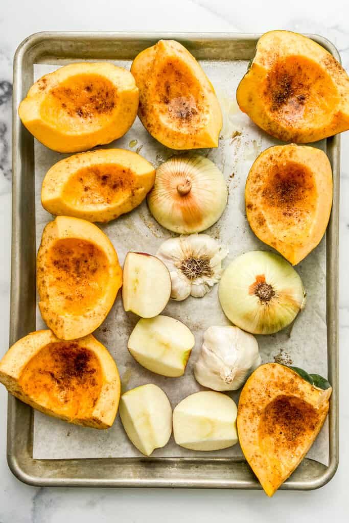 Quartered acorn squash topped with spices, apples, garlic, and an onion on a parchment lined baking sheet.