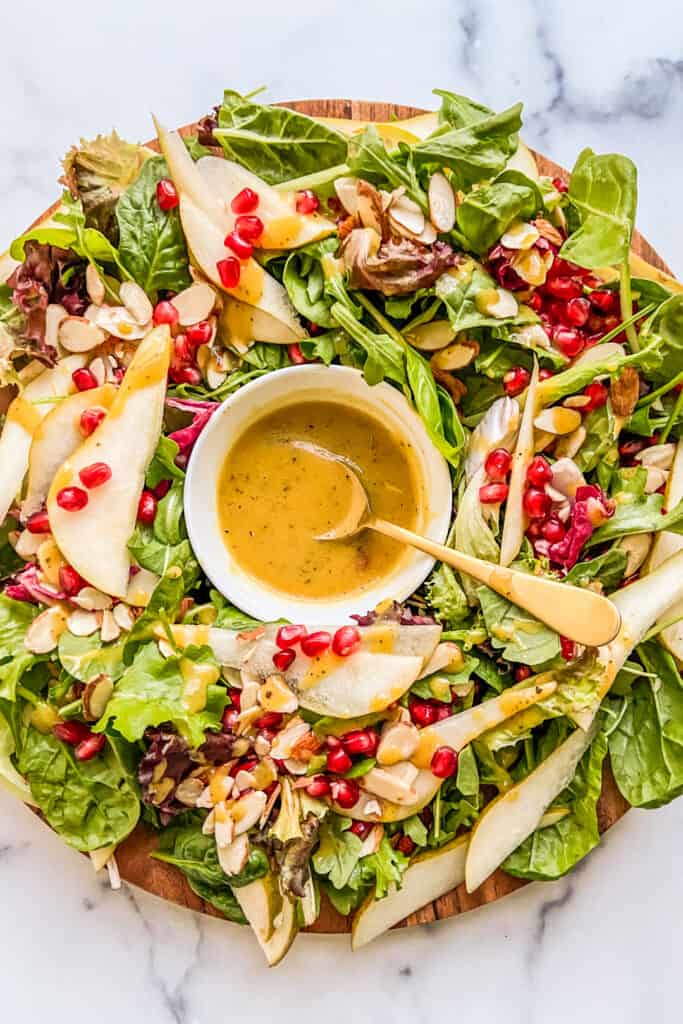 A holiday salad on a large wooden plate with a bowl of dijon dressing in the center.