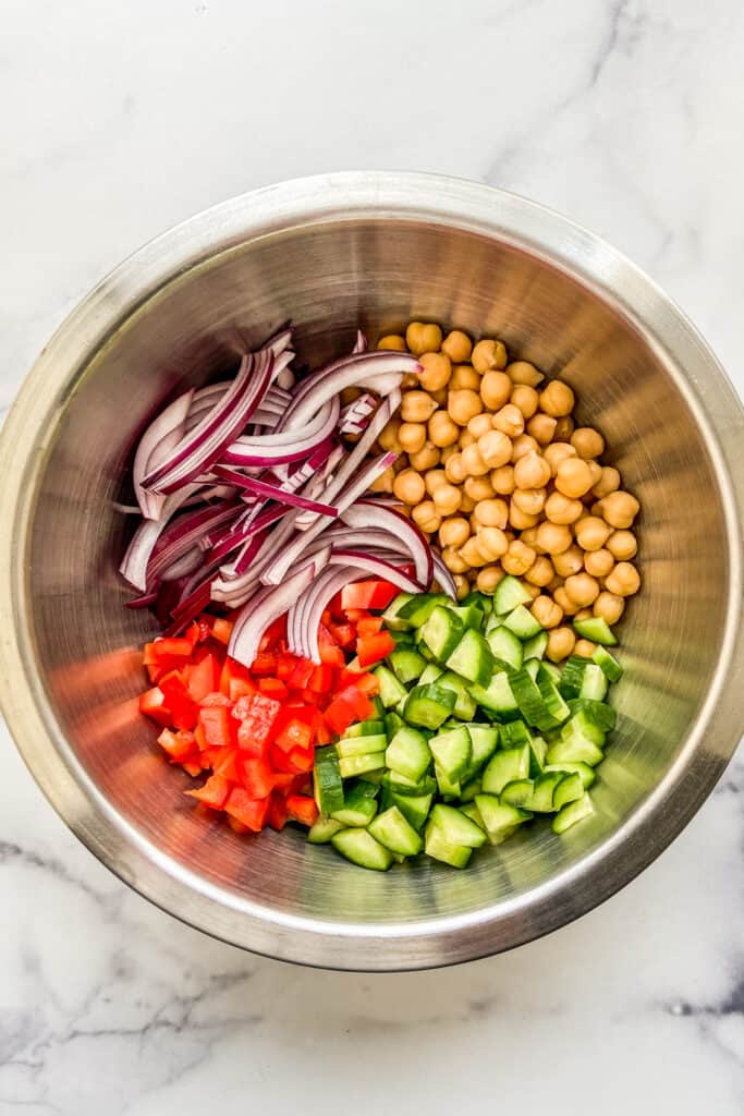 Chopped red onion, bell peppers, cucumbers, and chickpeas in a mixing bowl.