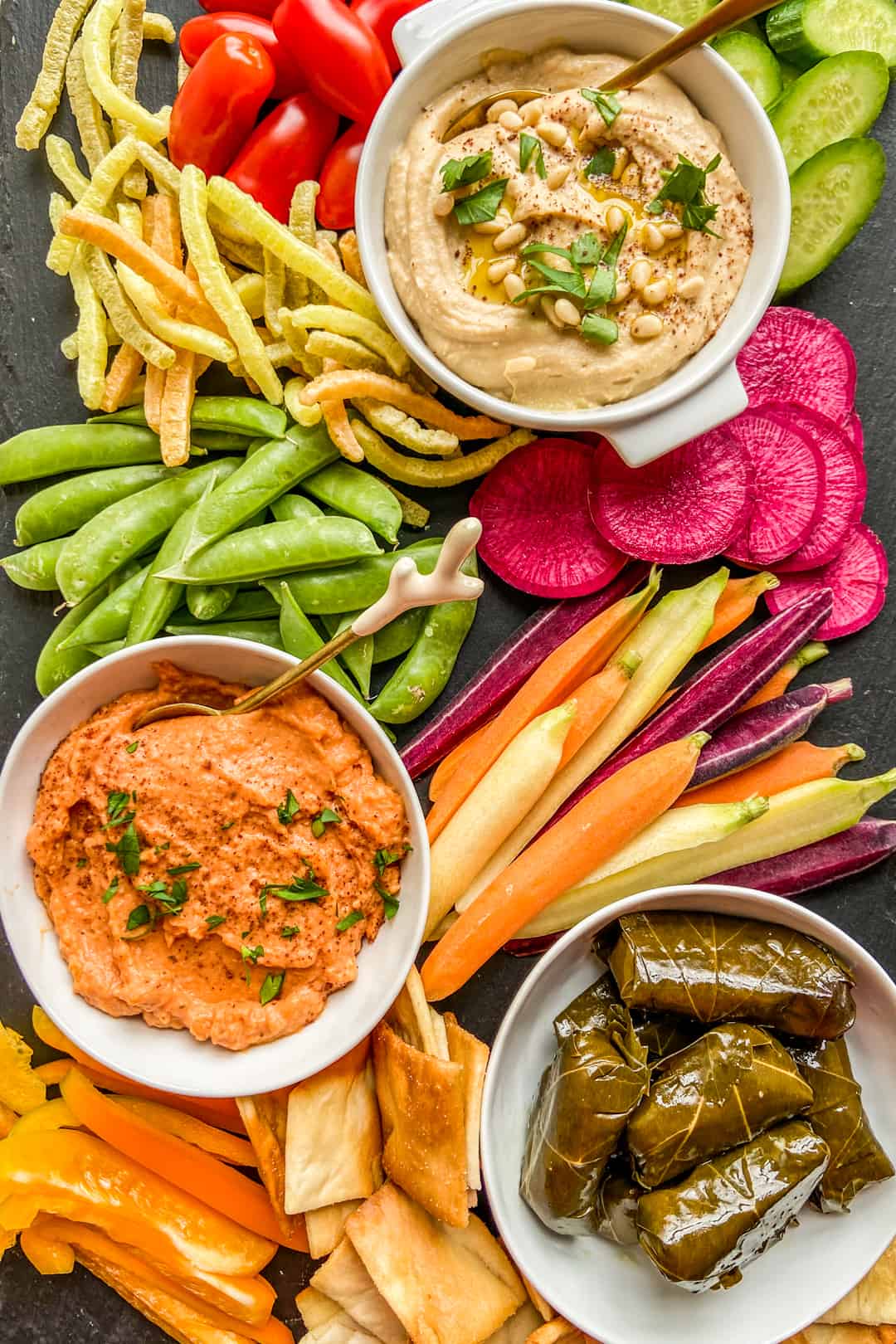 An overhead shot of a hummus board with hummus, grape leaves, cut vegetables, pita chips, and veggie straws.