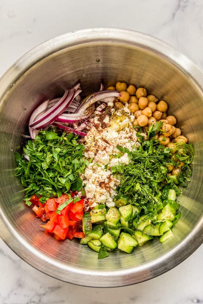 Chopped vegetables, chickpeas, feta cheese, fresh herbs, olive oil, and spices in a metal mixing bowl.