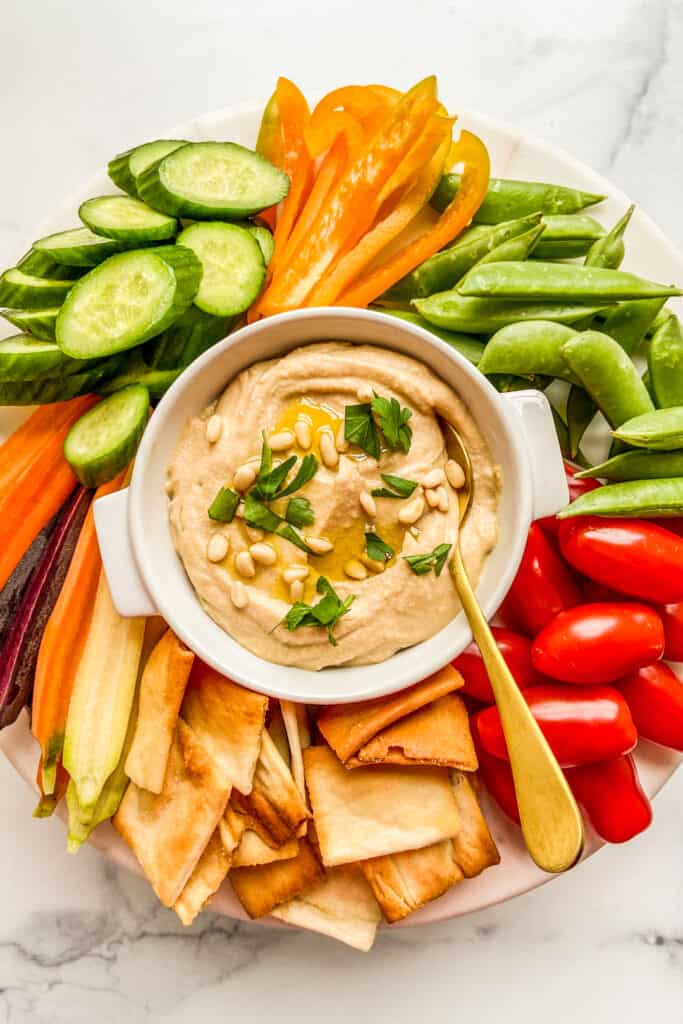 A bowl of hummus surrounded by pita chips and cut vegetables.