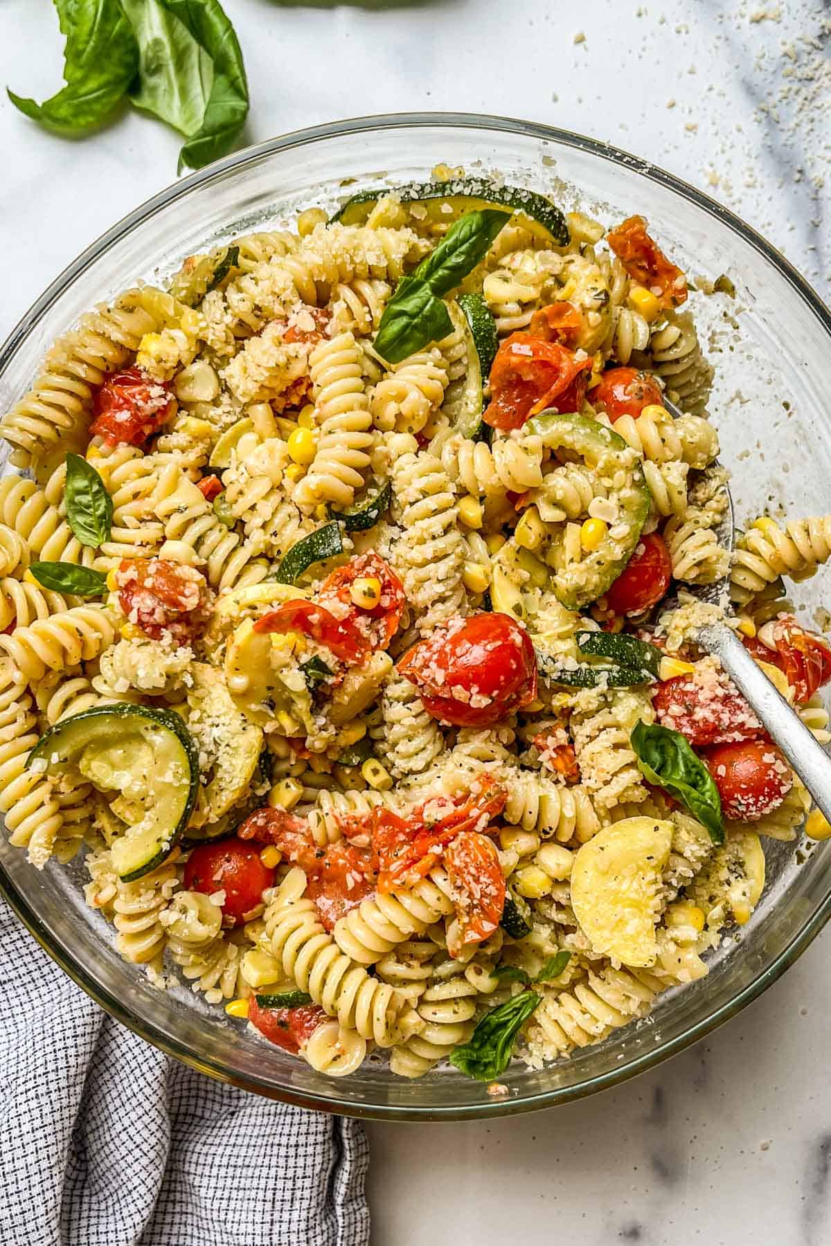 Pesto pasta salad with zucchini, summer squash, corn, and cherry tomatoes in a glass bowl with a silver spoon.