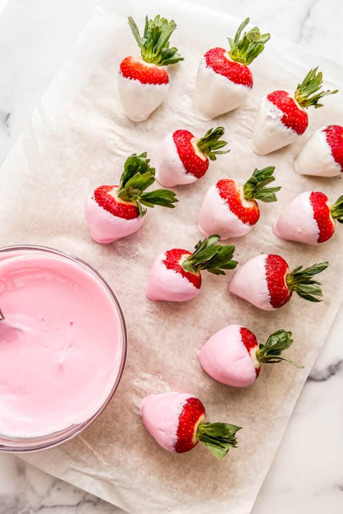 A bowl of pink yogurt and strawberries covered in yogurt on a piece of parchment paper.