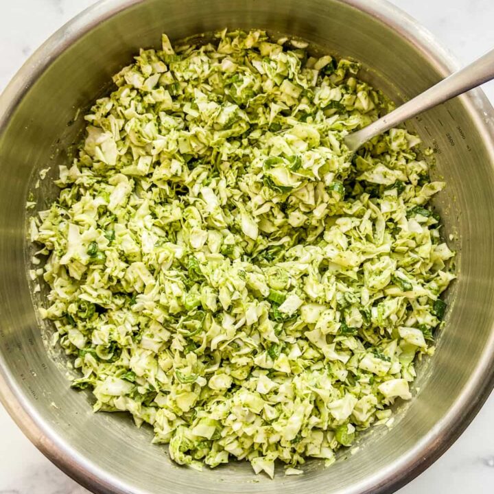 A green cabbage salad in a metal mixing bowl with a serving spoon.