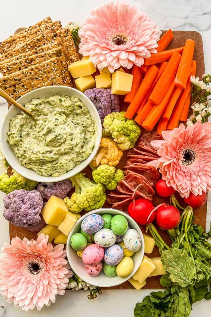 An Easter charcuterie board with flowers, dip, crackers, veggies, carrots, salami, and cheese.