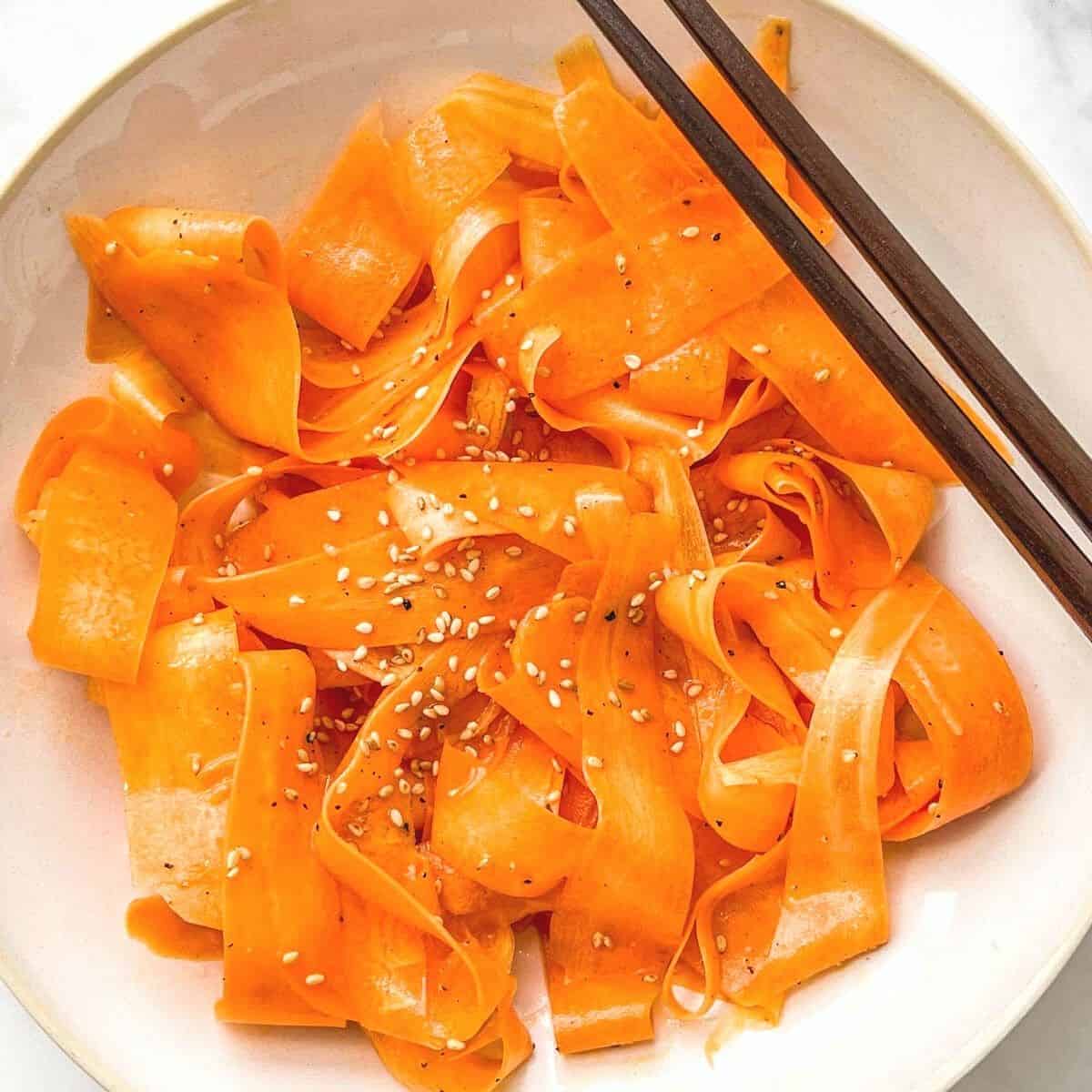 A shaved carrot salad in a white bowl with chopsticks.