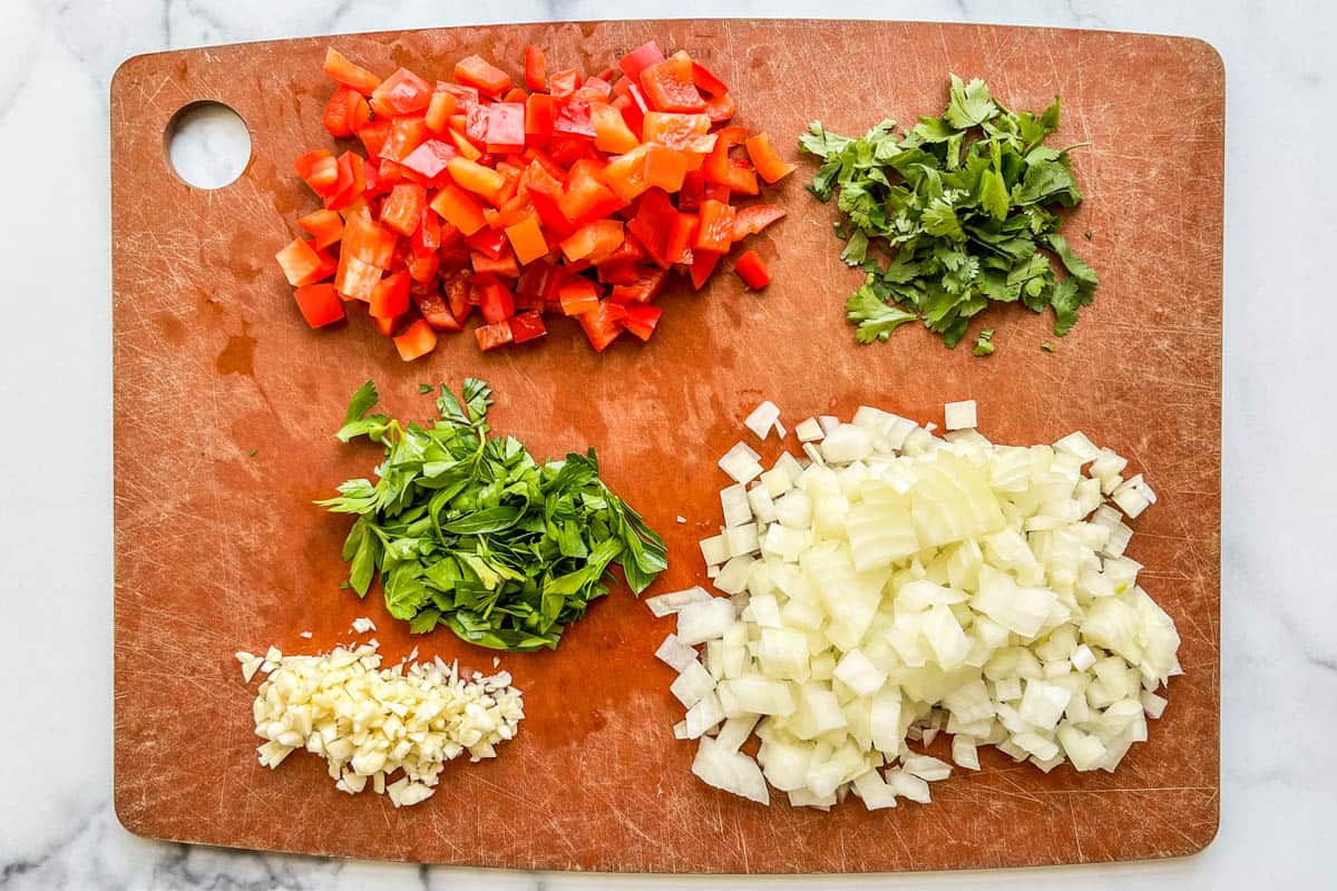 Chopped bell pepper, parsley, onion, basil, and garlic on a cutting board.