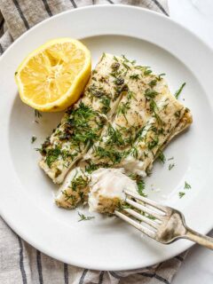 Pan seared halibut with dill, capers, and lemon on a white plate with a fork.