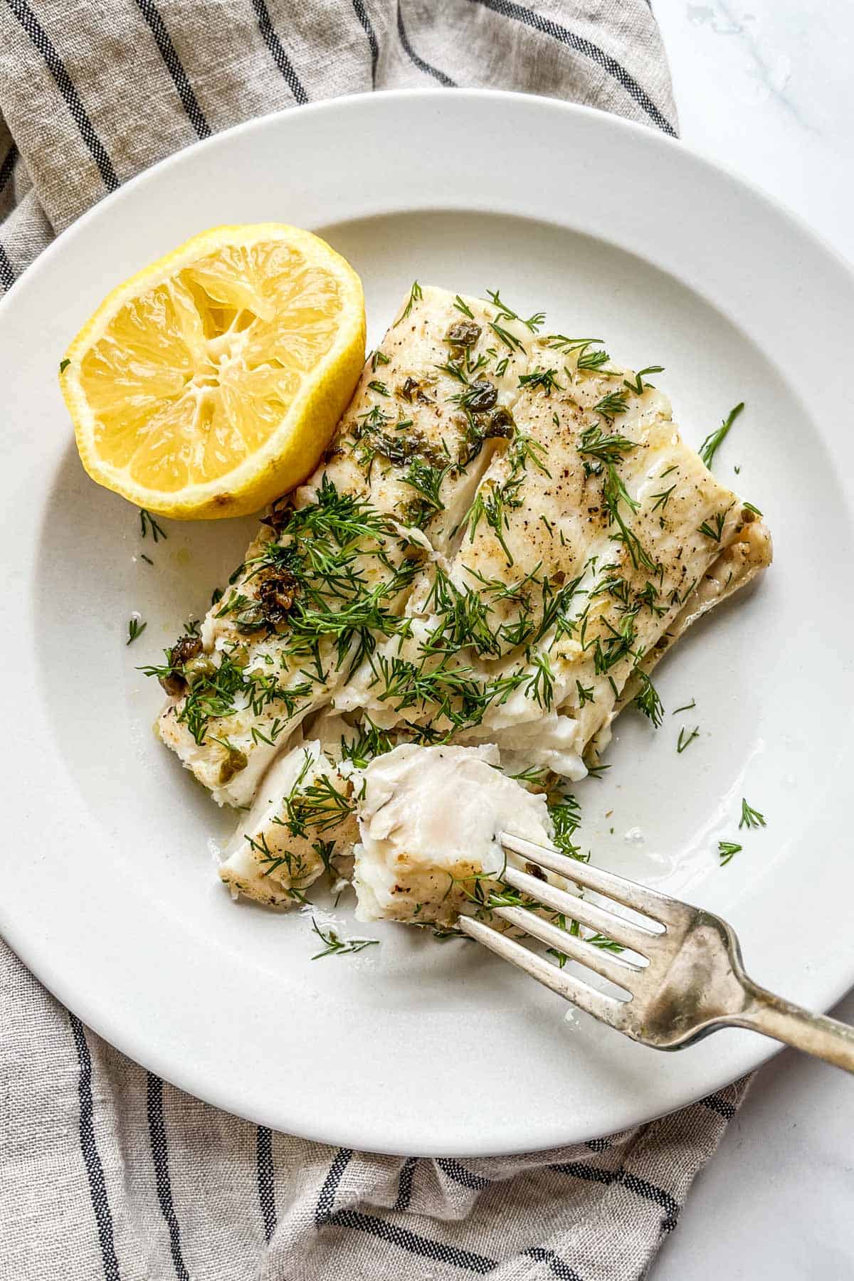 Pan seared halibut with dill, capers, and lemon on a white plate with a fork.