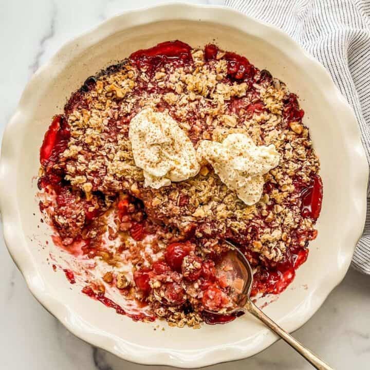 Sour cherry crisp in a white pie plate, topped with dollops of whipped cream.