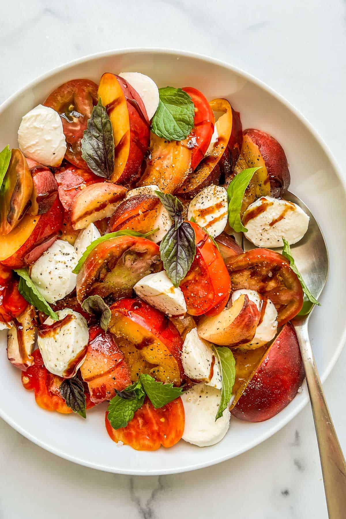 A stone fruit and heirloom tomato salad in a bowl with a large spoon.