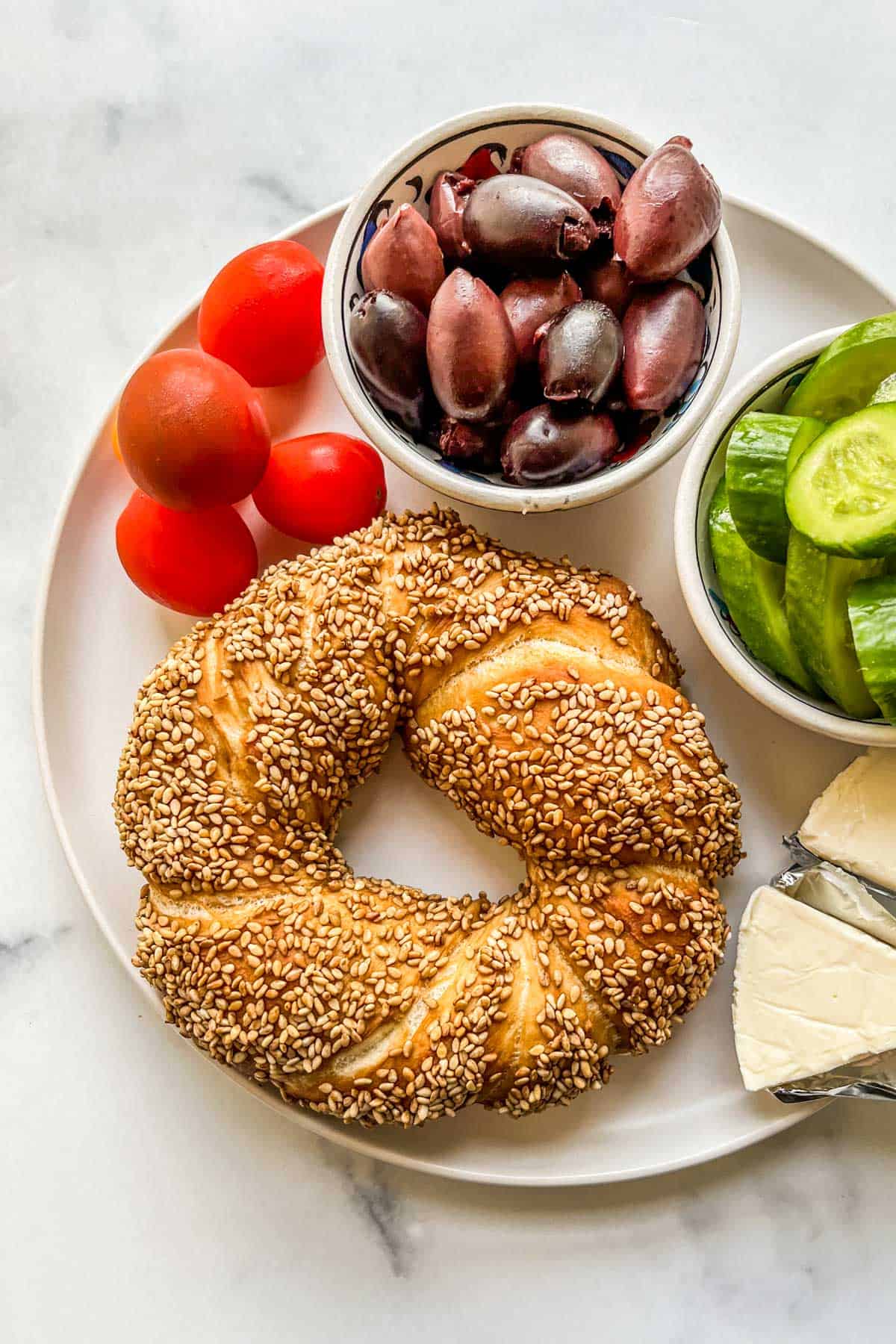 A Turkish simit bread on a plate with tomatoes, olives, cucumber, and cheese.