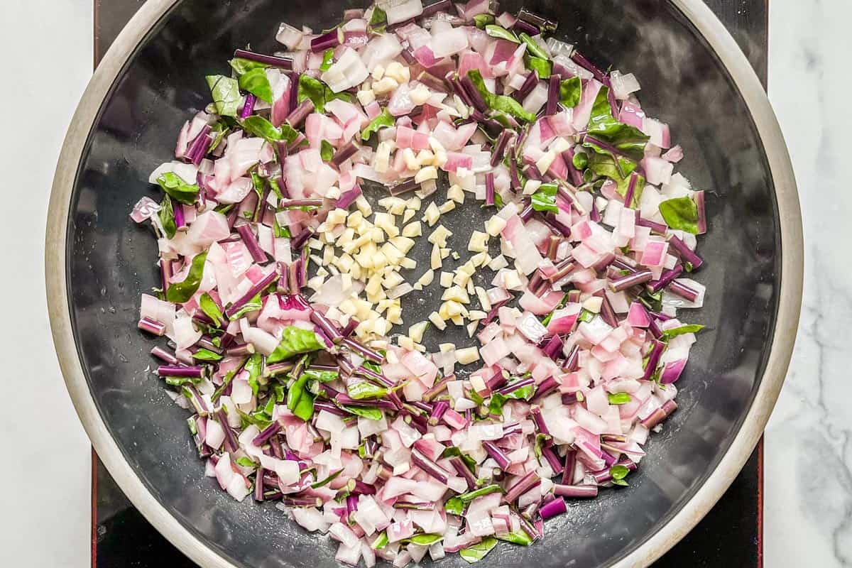 Minced garlic added to a skillet with kohlrabi stems and diced red onion.