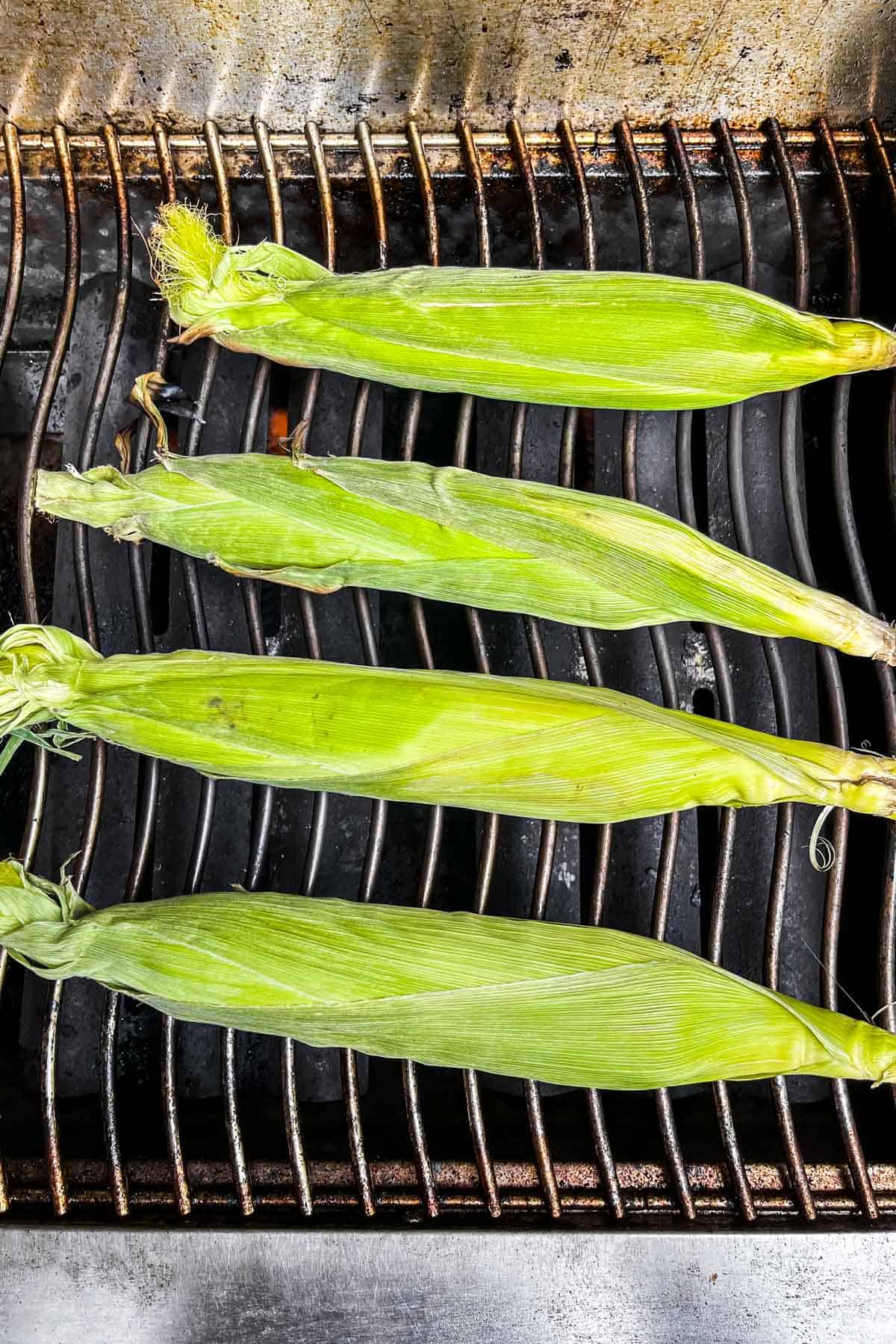 Four pieces of corn with green husks on a grill.