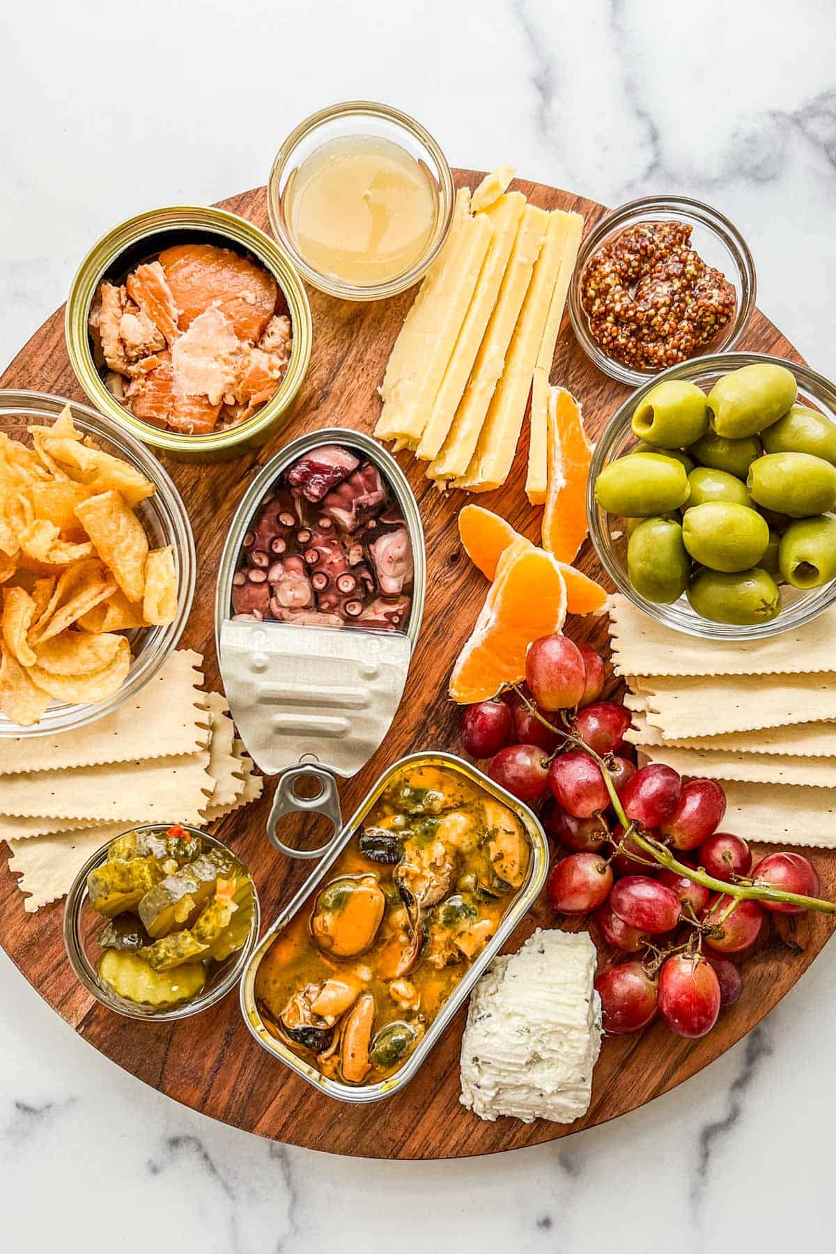 Several tinned fishes, crackers, cheese, and fruit on a wooden board.