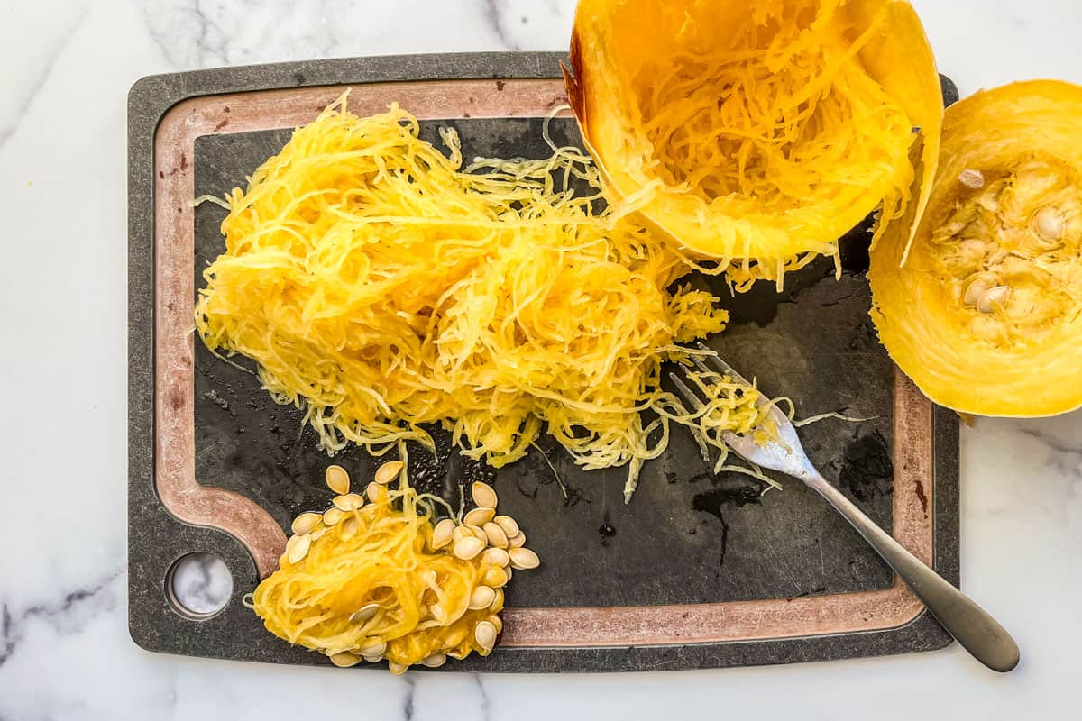 The noodle strands of half a spaghetti squash pulled out of the skin on a cutting board.