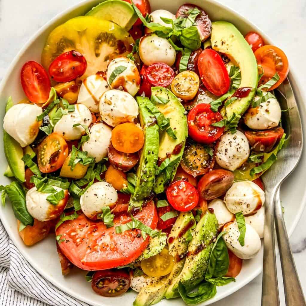 An avocado Caprese salad in a large white serving bowl with serving spoon and fork.