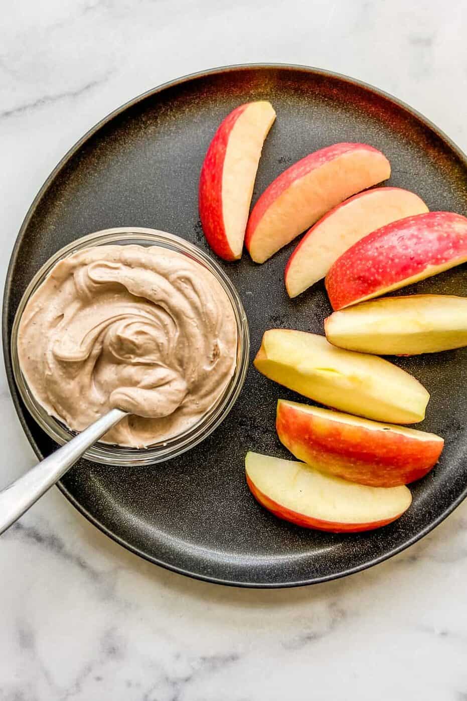 A small bowl of Greek yogurt dessert dip next to apple slices on a black plate.