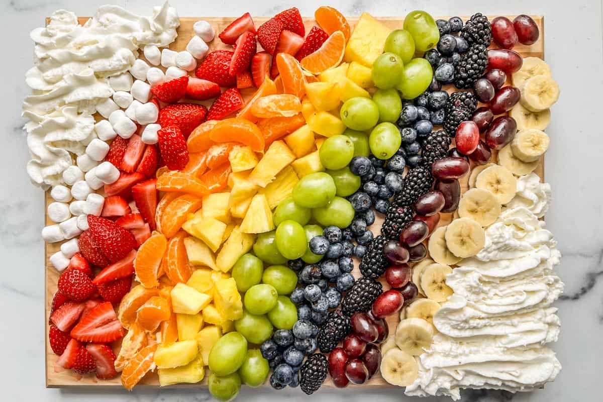 Rainbow fruit, whipped cream, and mini marshmallows on a cutting board.