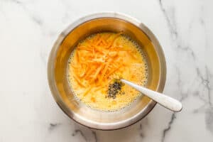 Eggs, milk, cheddar cheese, salt, and pepper being whisked in a metal bowl.