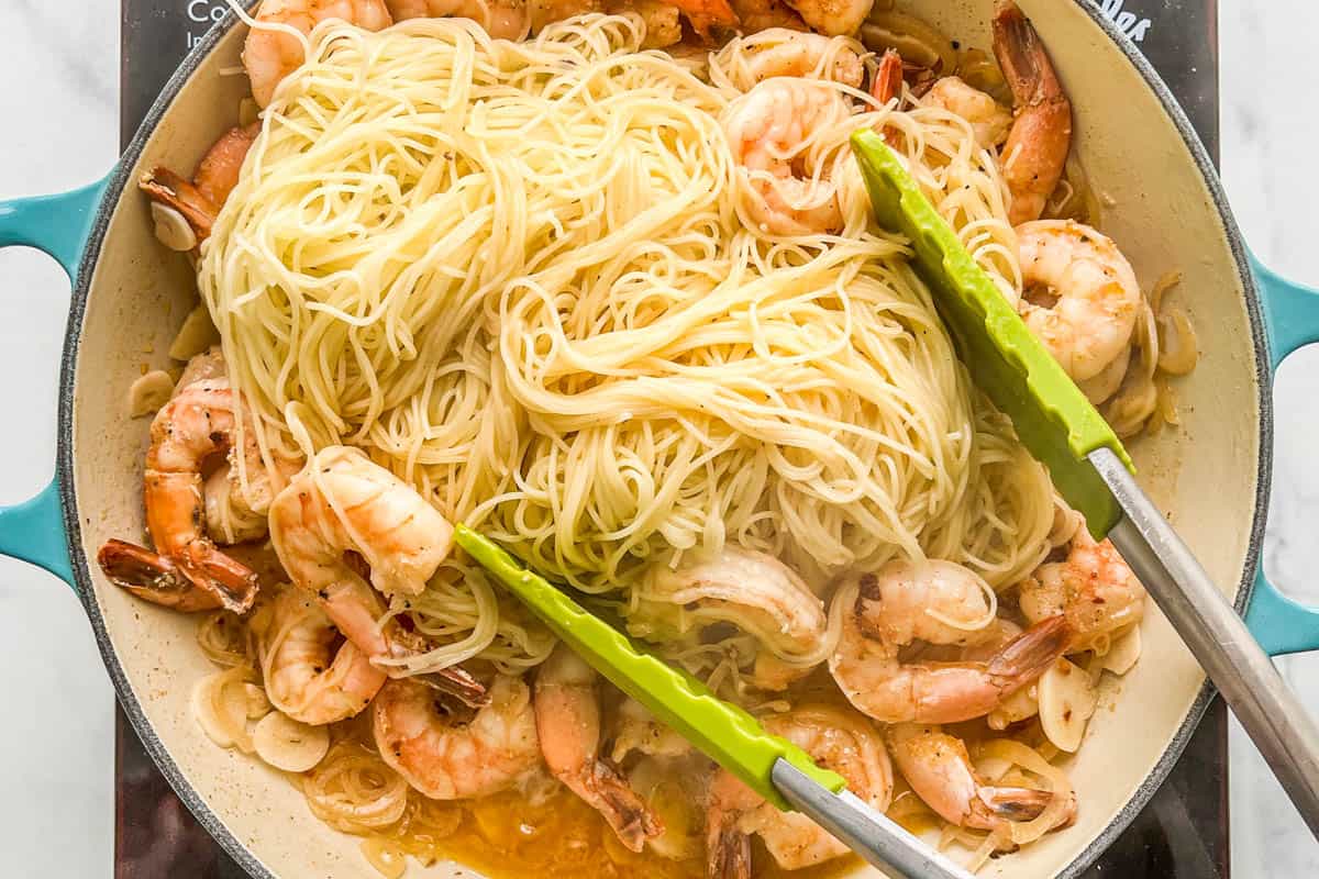 Angel hair pasta being added to a shrimp sauce in a skillet.