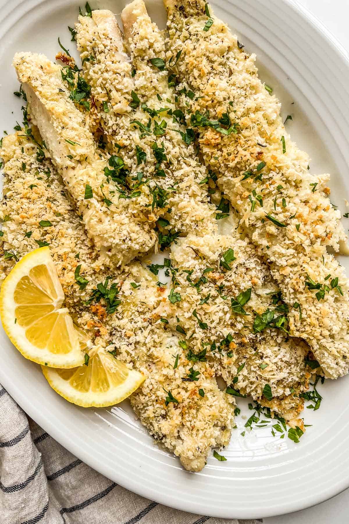 Baked pollock topped with parsley and served on a white platter with lemon slices.