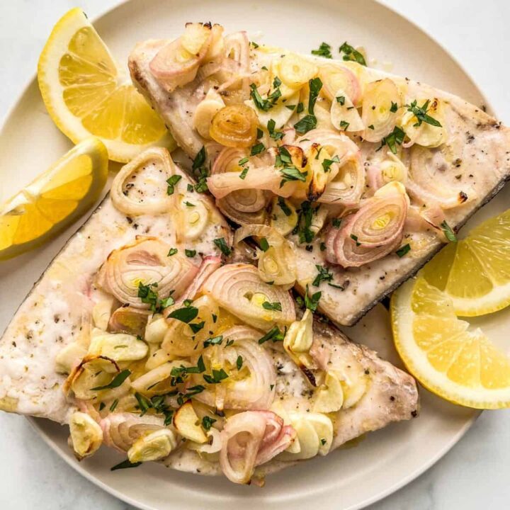 Two baked swordfish steaks on a white plate topped with lemon and shallots.
