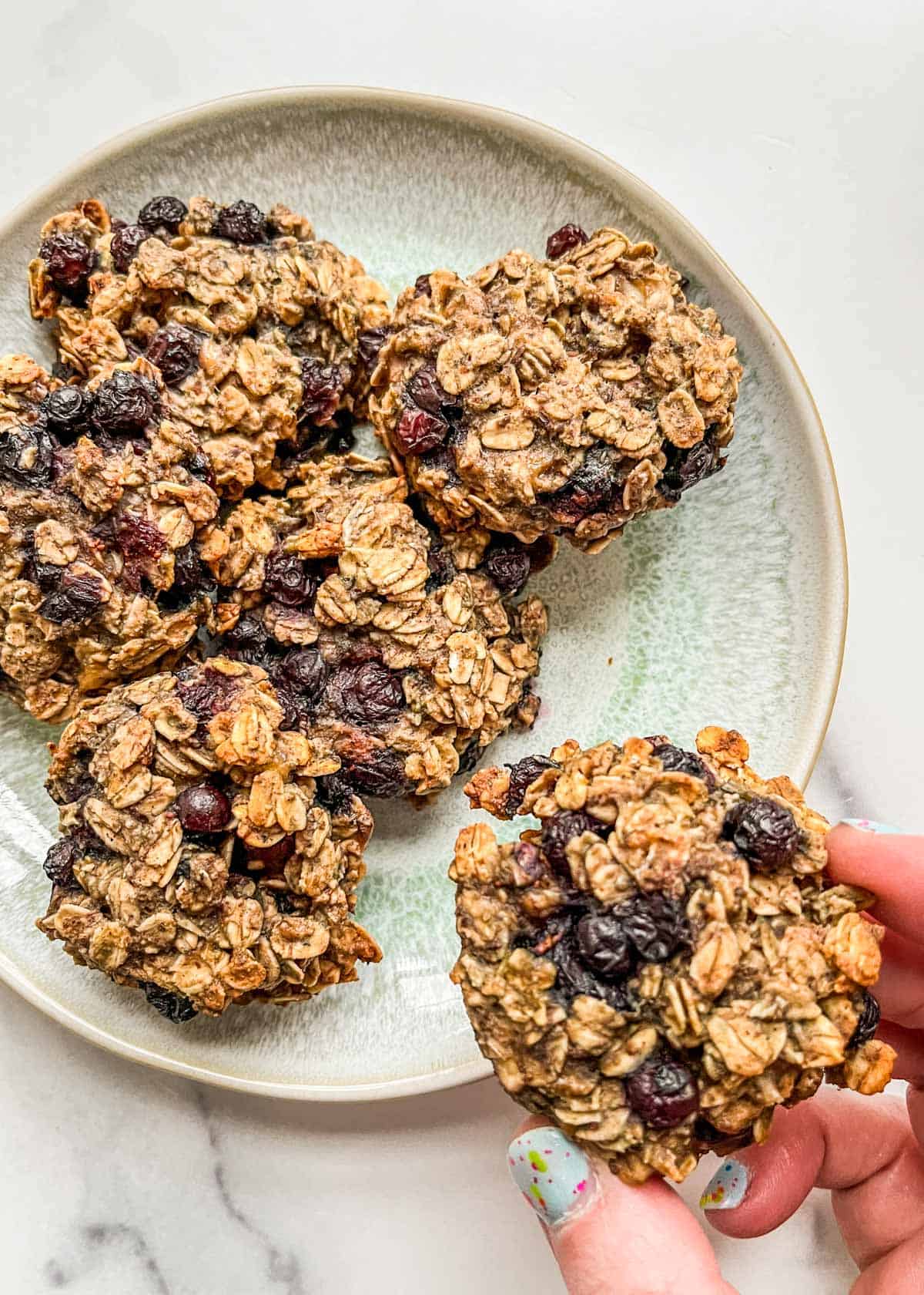 A hand holding one breakfast cookie next to a plate of cookies.